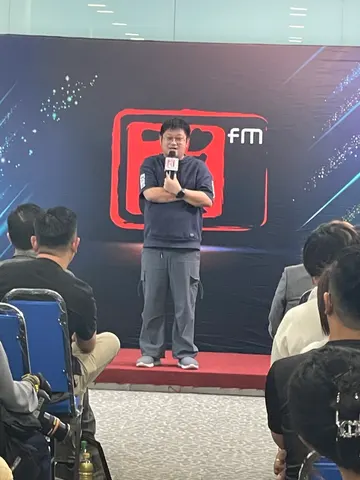 Mike bercakap menggunakan mikrofon di hadapan latar acara Ai FM.