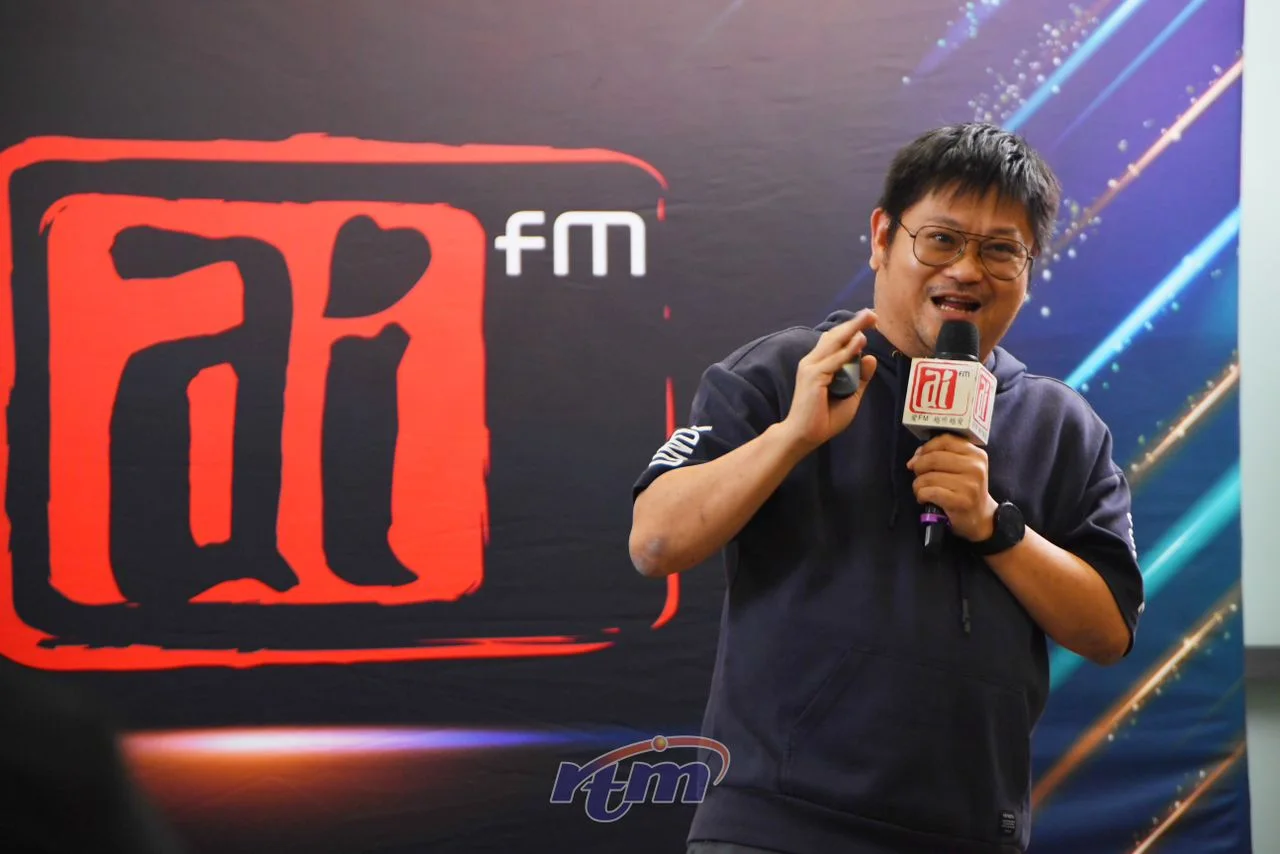 Mike bercakap menggunakan mikrofon di hadapan latar acara Ai FM.