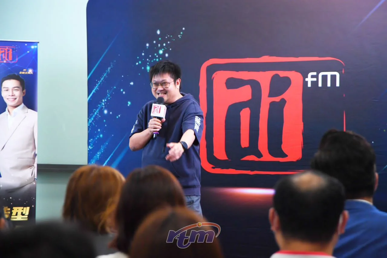 Mike bercakap menggunakan mikrofon di hadapan latar acara Ai FM.